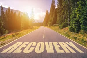 Image of a sunlit road through a wooded area, with the word "RECOVERY" imposed over it
